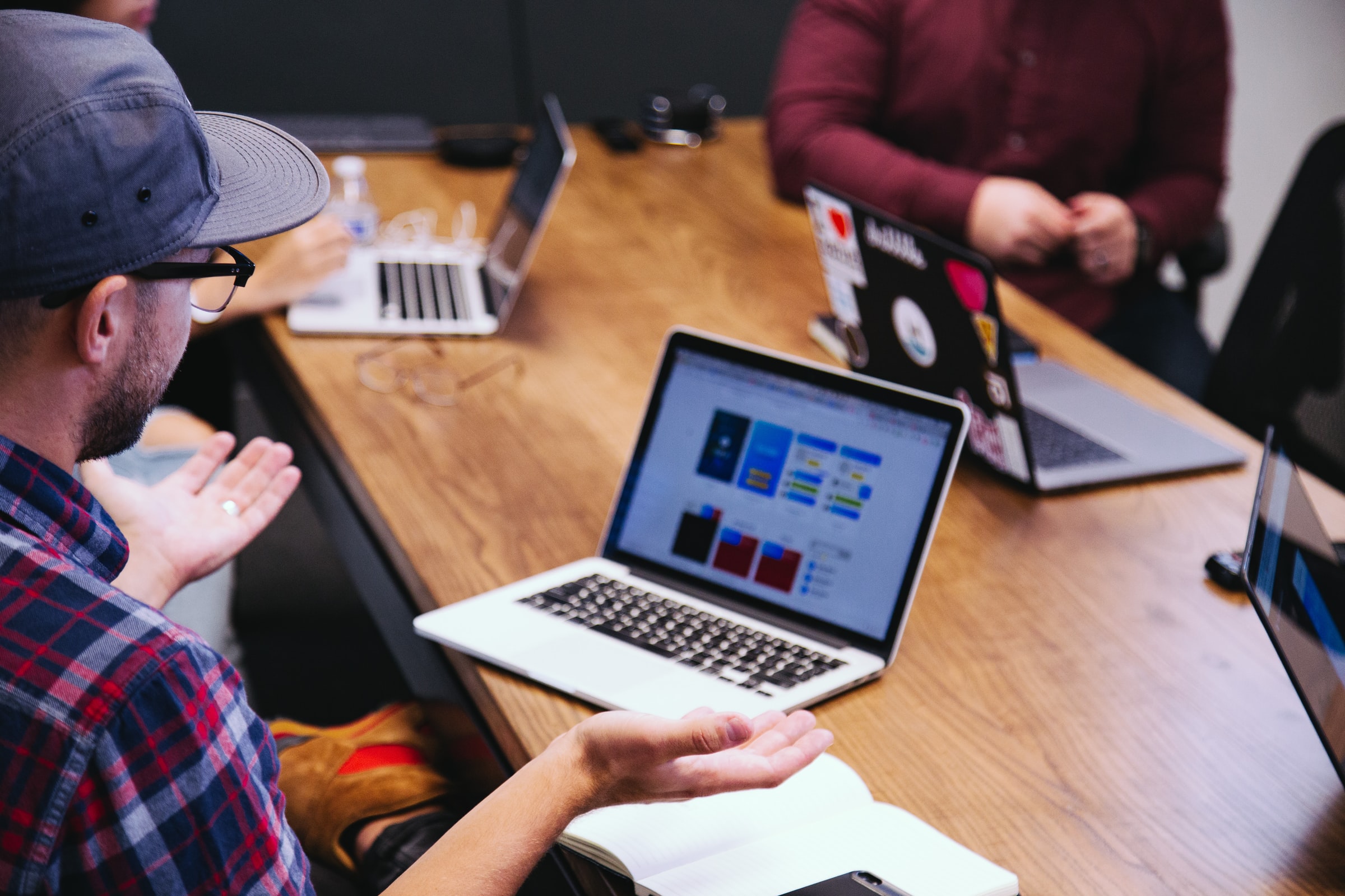 Team meeting in which multiple colleagues discuss in a table, where several computres are opened. One man moves hands displaying confusion